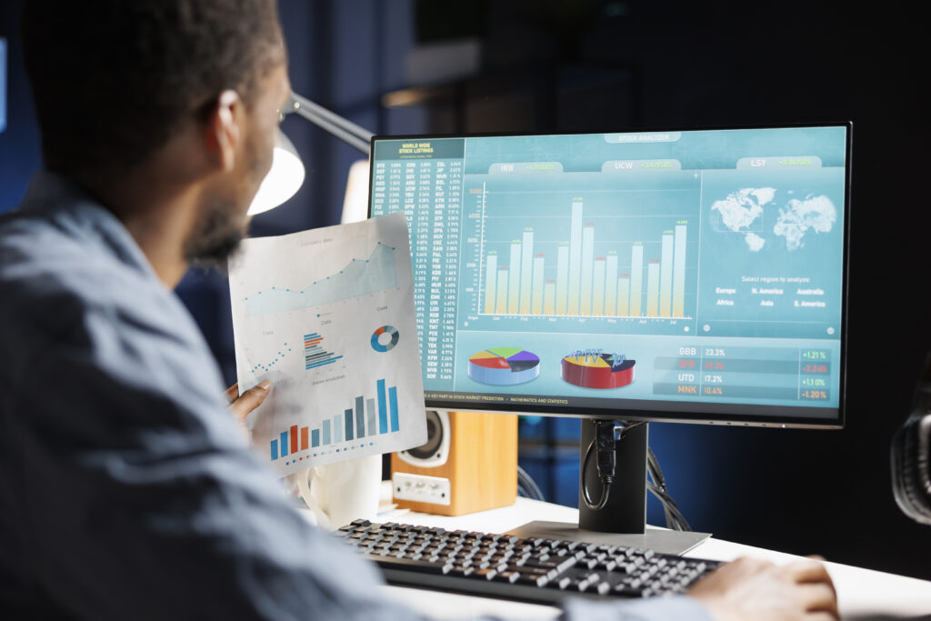 Vista desde atrás de un hombre analizando gráficos financieros complejos en un monitor de computadora y comparándolos con un reporte impreso en su mano. La pantalla muestra tablas de valores, gráficos de barras y un mapa mundial en tonos turquesa.