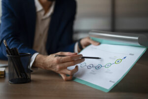 Primer plano de las manos de un ejecutivo en traje señalando con un lápiz un gráfico de línea ascendente en una carpeta. El gráfico muestra una secuencia de iconos financieros y una flecha azul que indica crecimiento y éxito empresarial.