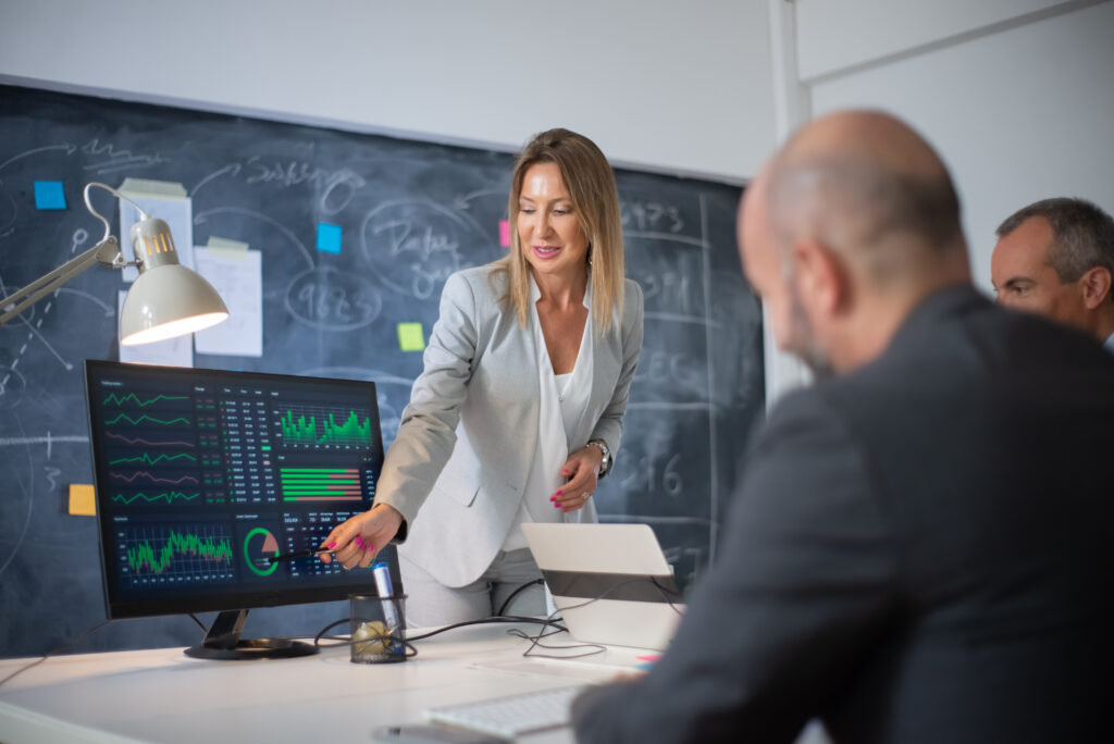 presentacion-indicadores-clave-rendimiento-inteligencia-de-negocios-bi Una experta señala un monitor que muestra dashboards financieros detallados frente a su equipo, facilitando la toma de decisiones mediante herramientas de inteligencia de negocios BI en un entorno de oficina.
