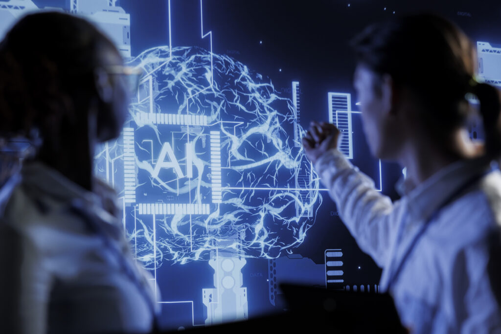 cientificos-analizando-cerebro-digital Dos profesionales de perfil, desenfocados en primer plano, observan una gran pantalla azul brillante. En la pantalla se muestra una representación digital de un cerebro humano integrado con circuitos electrónicos y las siglas "AI" en el centro, simbolizando redes neuronales e inteligencia artificial.
